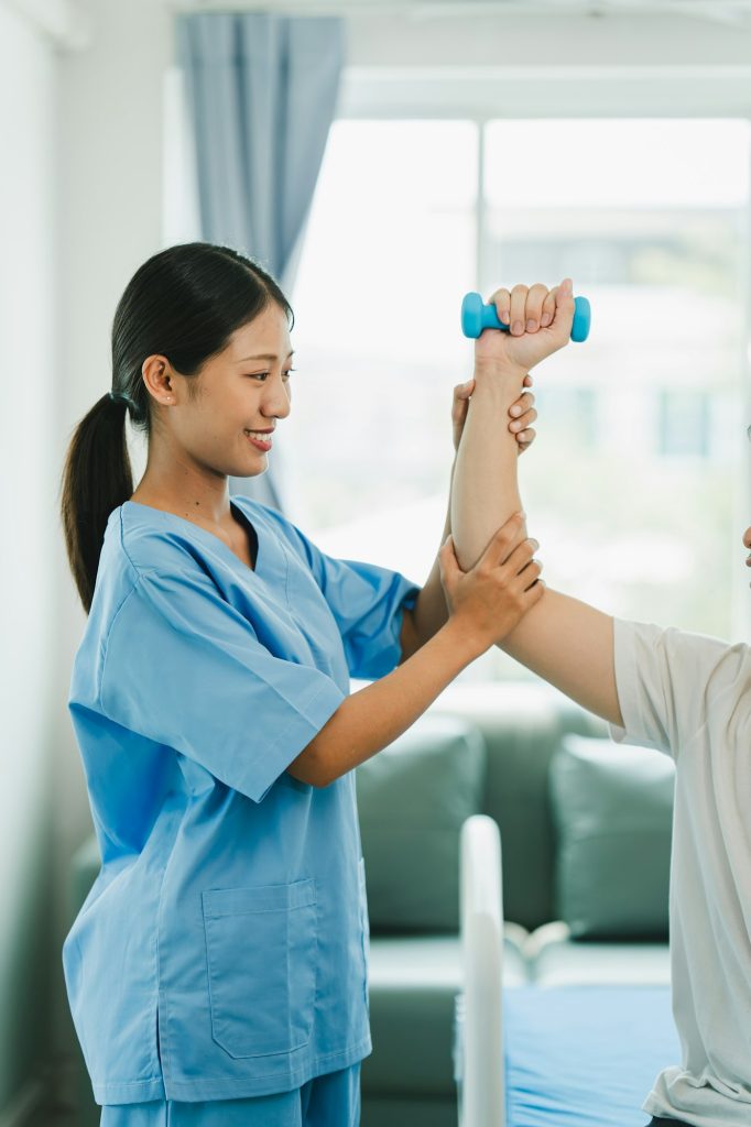 Physical therapist giving exercise by maintaining dumbbells on the arms and shoulders of a male pati